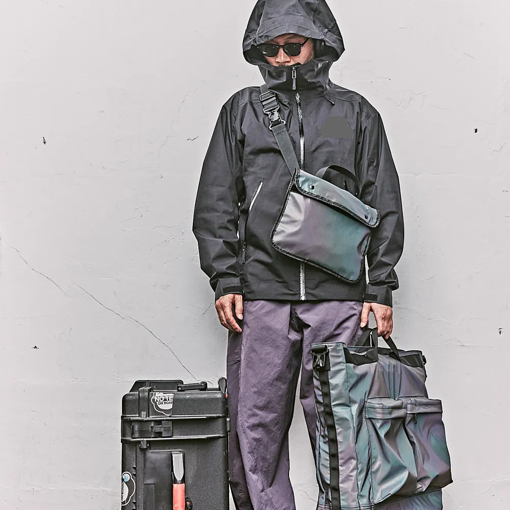 Ein Mann mit 2 Taschen, eine als Crossbody Bag und andere in der Hand als Henkeltasche Tote Bag, daneben ein schwarzer Koffer, kontrastreich dunkel vor weißem Hintergrund