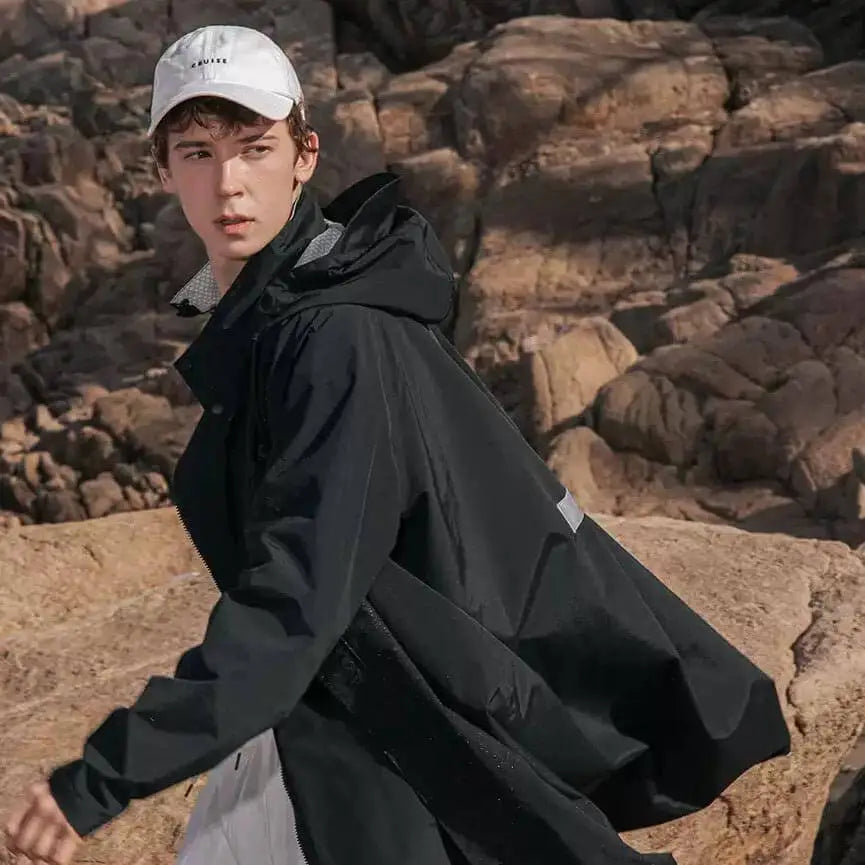 Ein Mann trägt den schwarzen oversized Regenmantel und geht. Er schaut nach hinten. Im Hintergrund braune Felsen.