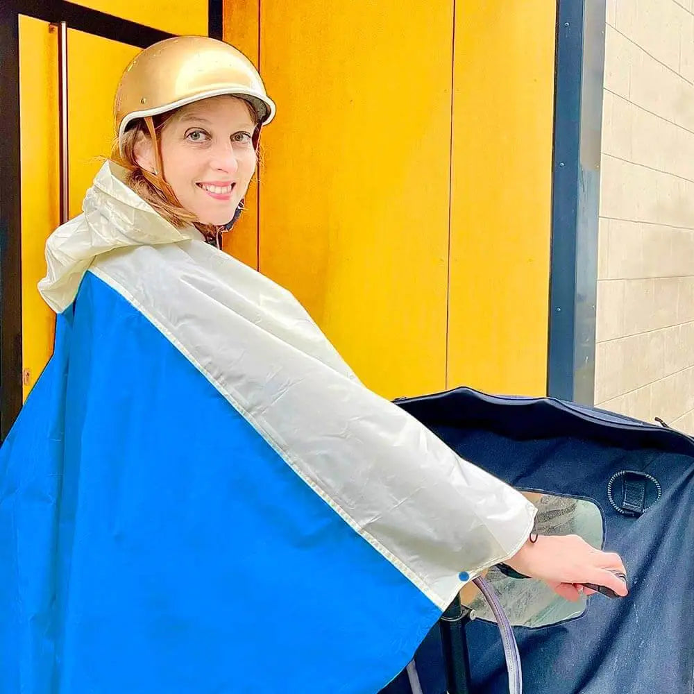 blau-weißer Regenponcho mit Lastenrad an einer Frau mit Fahrradhelm vor einer gelben Wand Hauseingang