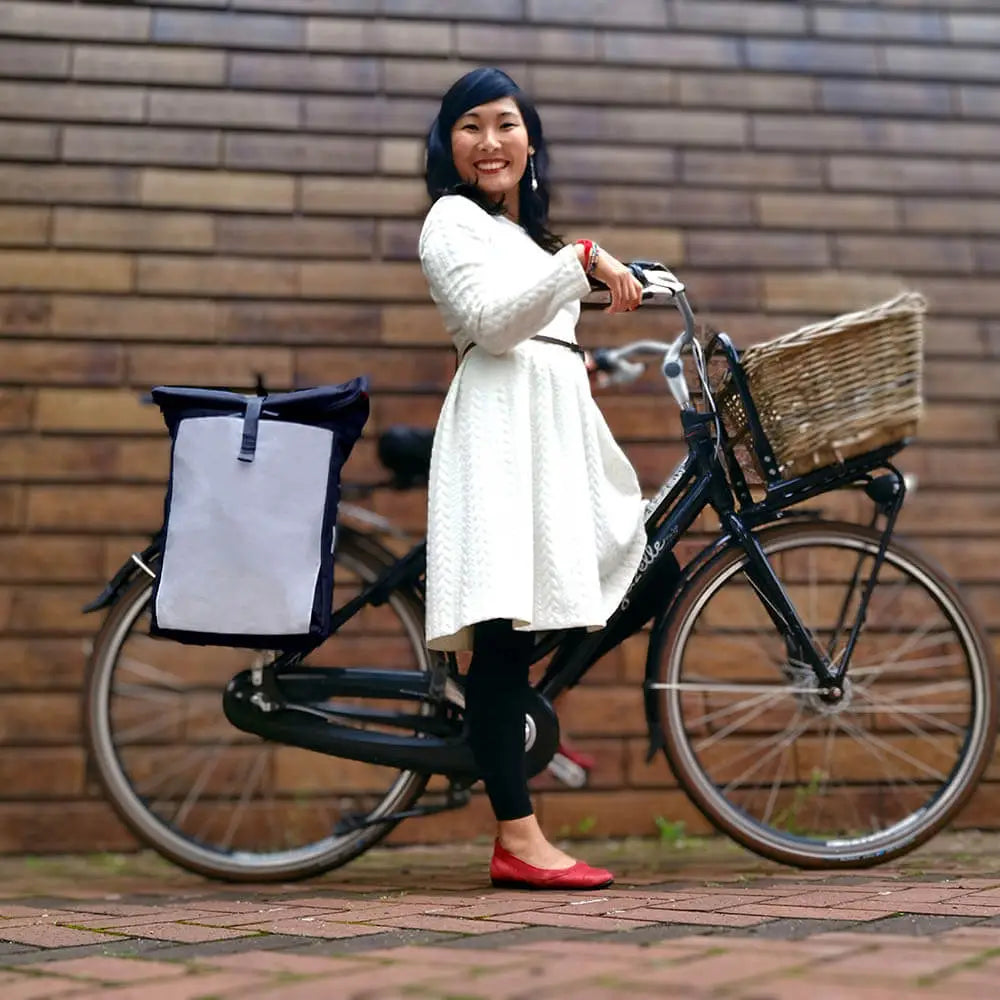 Frau auf dem schwarzen Fahrrad Hollandrad in einem schicken weißen Kleid in der Stadt vor einer Brick Wand mit der schicken Bomence Urban Create reflektierende Fahrradtasche und Rucksack 2-in-1. Der Fahrradrucksack ist schwarz grau und hat einen Rolltop Verschluss, für urbane Pendler.