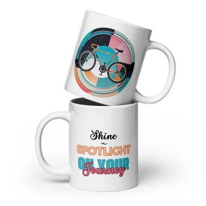 Weiße glänzende Kaffeetasse mit 20 oz Fassungsvermögen, schlichte und elegante Vorderansicht. Weiße Kaffeetasse mit einem bunten Fahrradmotiv auf der einen Seite und dem inspirierenden Spruch „Shine a Spotlight on Your Journey“ auf der anderen Seite.