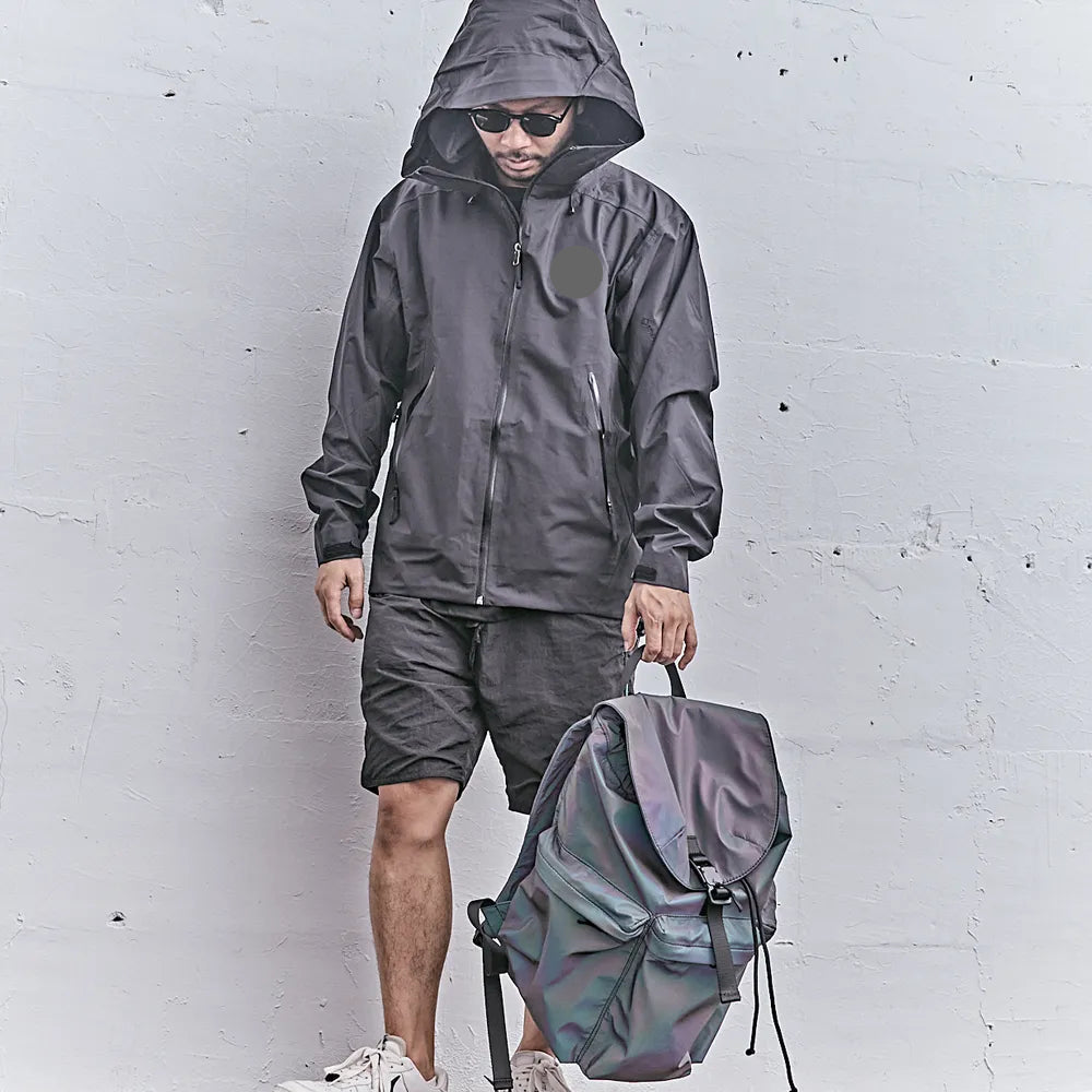 cooler Mann mit dunkler Jacke und Kapuze und Sonnenbrille und shorts mit Rucksack aus reflektierendem Material in der Hand hängend