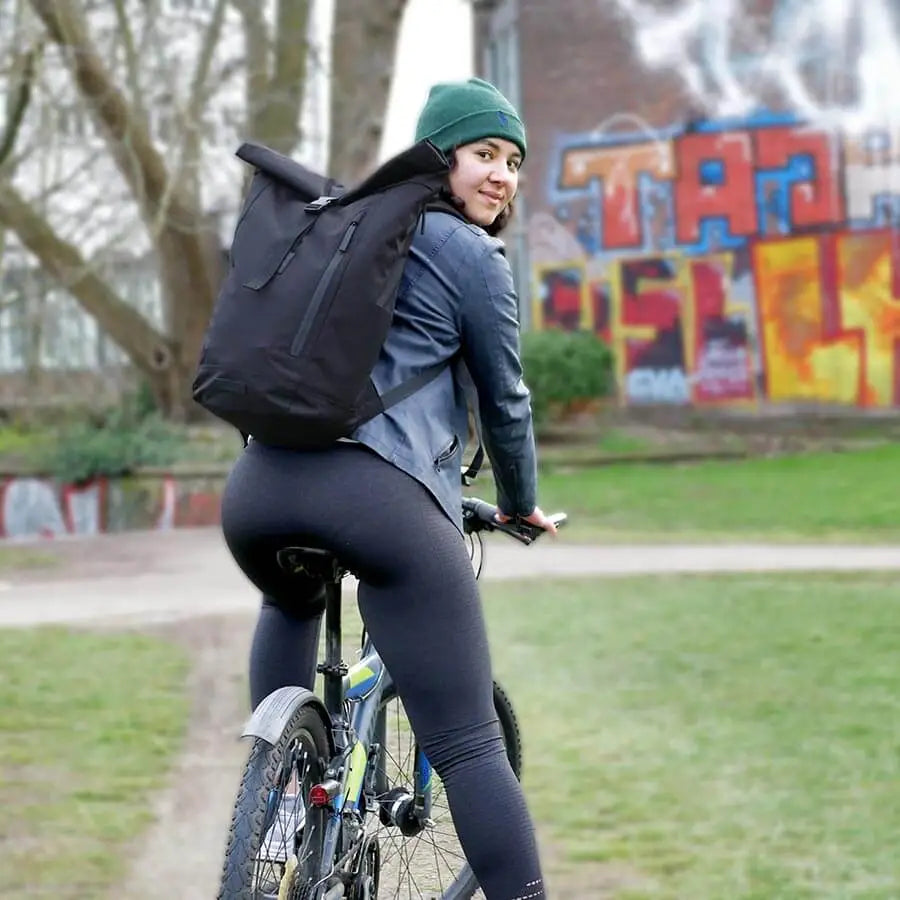 Frau auf dem Fahrrad blickt zurück in die Kamera mit einem schwarzen Bomence Rolltop Rucksack auf dem Rücken. Hintergrund urban outdoor mit Graffiti.