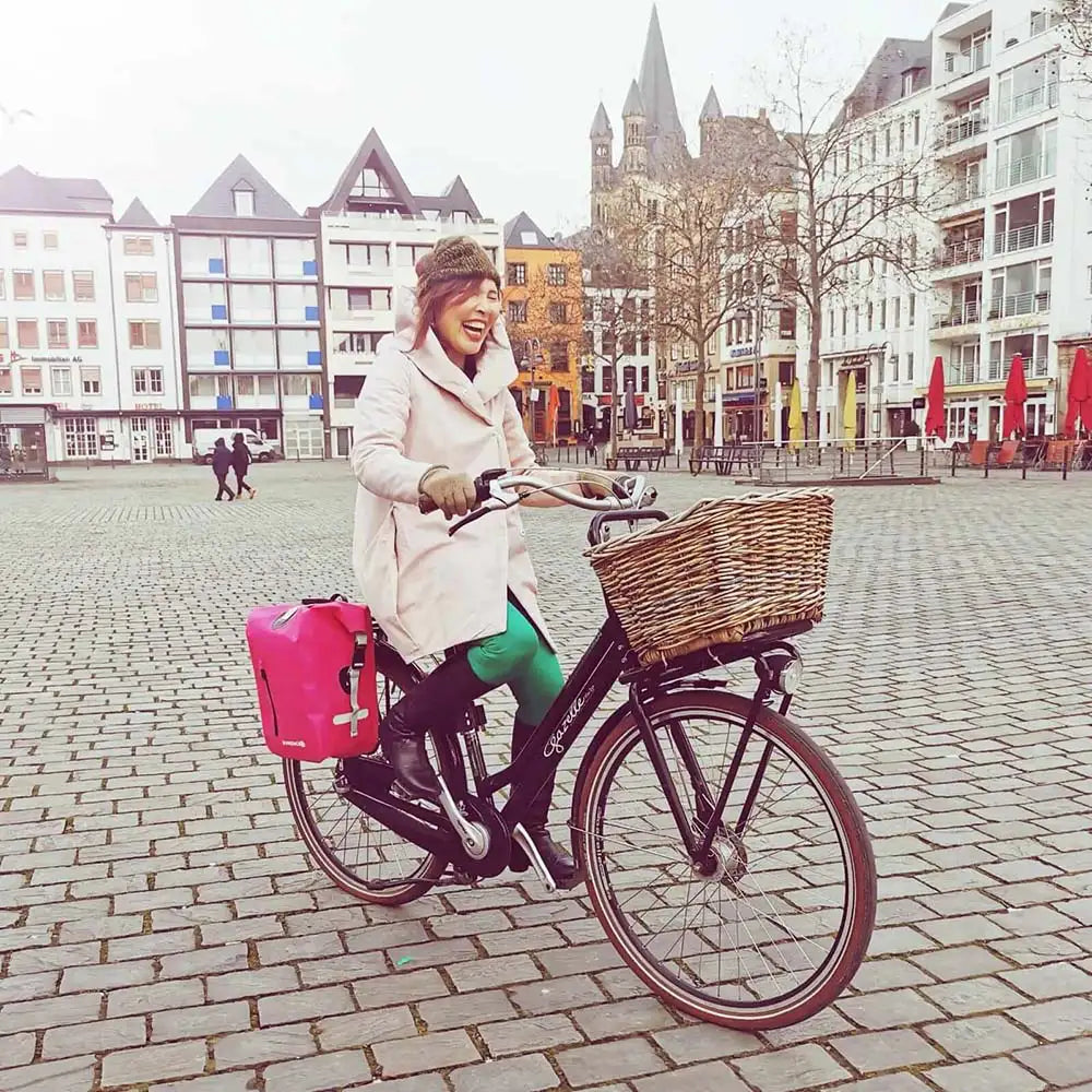 Eine Frau fährt Fahrrad in Köln mit einer pinken Fahrradtasche am Gepäckträger des Hollandrads. Sie ist glücklich und lacht.