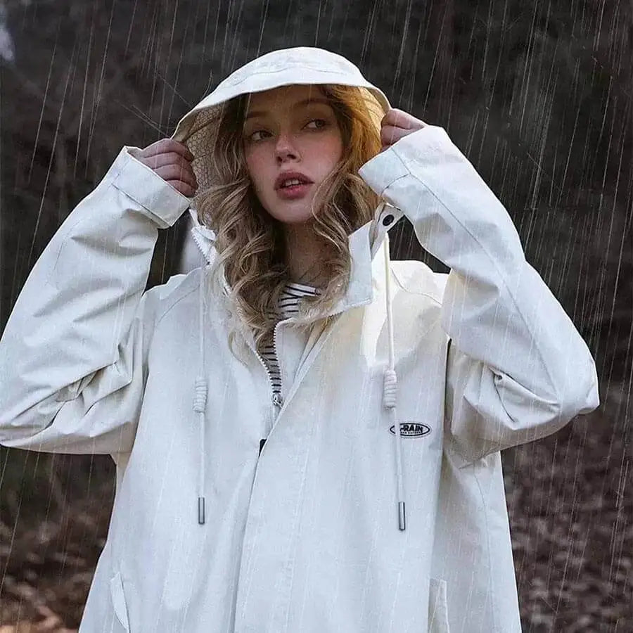 Weißer Regenmantel oversize lang mit kapuze und Reiverschluss Frau trägt den Mantel zieht die Kapuze auf und schaut nach rechts. Es regnet.