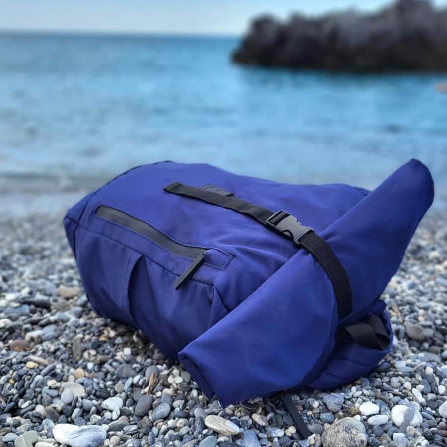 Blauer Rucksack mit Rolltop liegt verschlossen an einem Strand. Prägnant ist der Verschluss und die seitliche Tasche mit Reisverschluss.