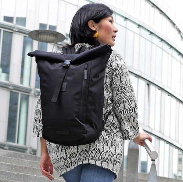 Schwarzer Rolltop Rucksack recycelt an einer asiatischen Frau tragend mit schwarz-weiß gemusterter Jacke schickes outfit vor einem Glasgebäude die Treppe empor steigend seitlich schauend mit Blick in die Ferne.