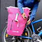 Video zeigt Packmöglichkeiten der rosa Roll-top Tasche und das leichte Anbringen am Fahrrad. Tasche verfügt über Quick-Lock Mechanismus. Rosa Radtasche wird aus verschiedenen Perspektiven gezeigt. Frau öffnen und schließt Tasche auf verschieden Arten. Die Frau trägt die Tasche zuerst als Umhängetasche und anschließend als Fahrradtasche.