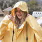Frau mit Blonden Haaren in einem gelben weiten Regenmantel, beide Hände ziehen die Kapuze auf, der Blick lächelnd und nach unten gerichtet. Bildausschnitt von Kopf bis Brust und Outdoor. Im Hintergrund Natur.