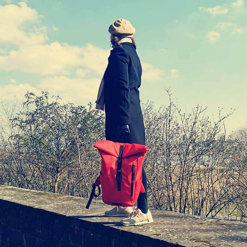 Eine Frau im schwarzen Mantel steht auf einer Mauer und schaut in die Ferne im Hintergrund winterliche Baumkronen aus Ästen und ein heller blauer Himmel mit Wolken mit Lederhandschuhen hält sie mit einer Hand den roten Rolltop Rucksack am Griff fest, sodass er über dem Boden schwebt nostalgischer Charakter des Bildes